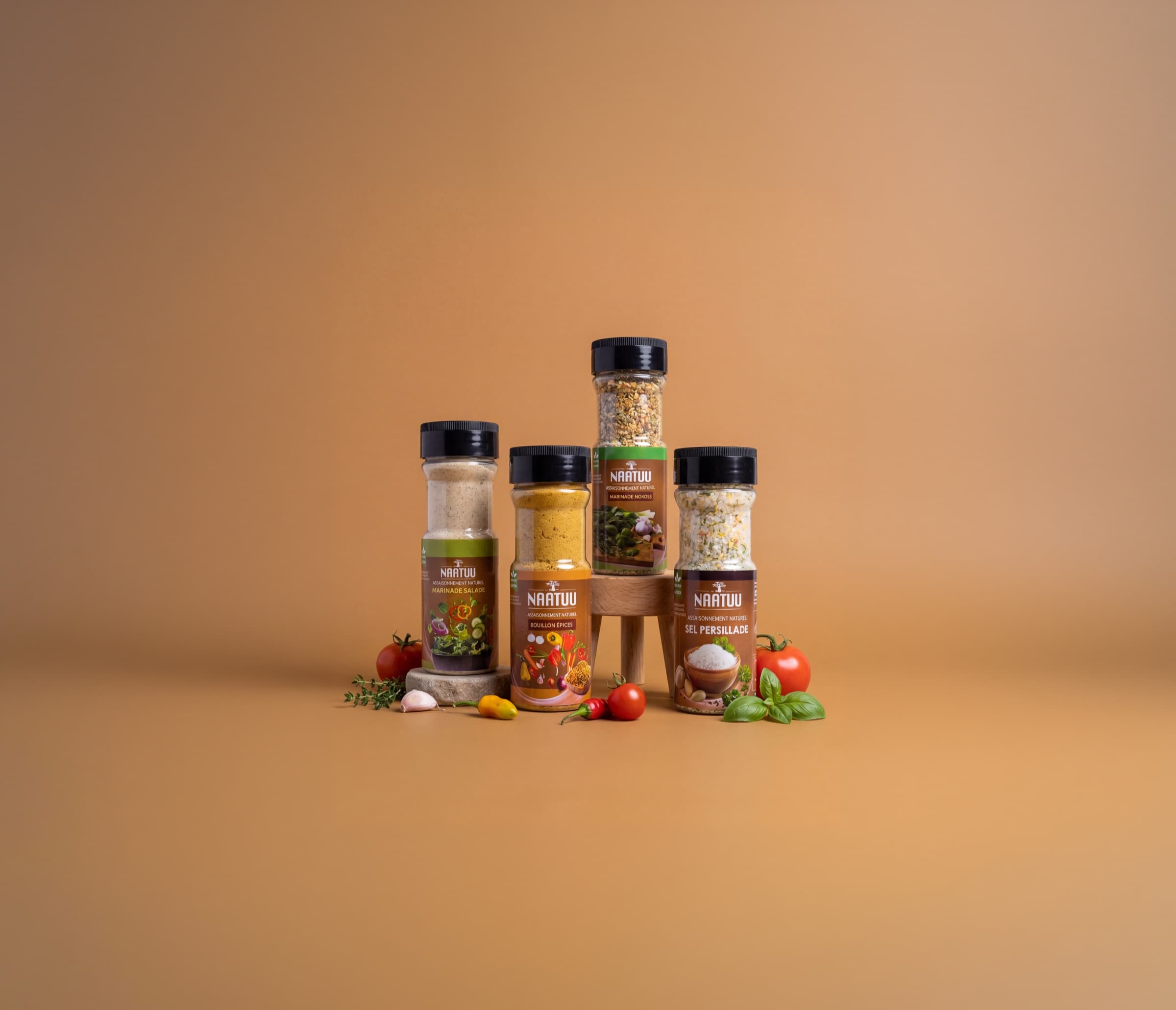Les quatre assaisonnements NAATUU, Marinade Salade, Bouillon Épices, Marinade Nokoss et Sel Persillade, entourés de tomates, d'ail, de piment et de basilic.